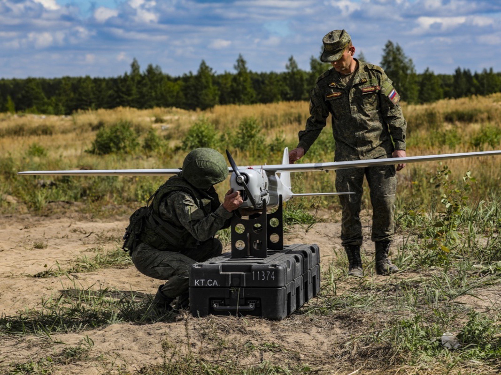 Небольшой беспилотник на подставке поверх транспортного кейса; один из двух военнослужащих проверяет аппарат, второй стоит рядом